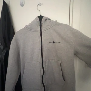 Grå hoodie från Sail Racing med dragkedja - Grå hoodie från Sail Racing med svart dragkedja och svart  logga på bröstet. Tröjan har huva, kängurufickor och är långärmad. Perfekt för dig som gillar en enkel och clean stil. Vissa av bokstäverna på loggan har försvunnit därav det billigs priset