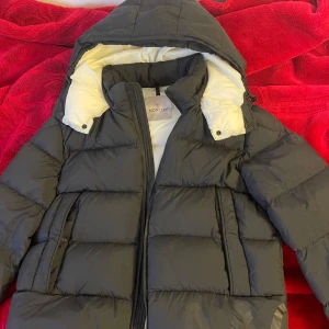 Svart Moncler dunjacka med huva - Snygg svart dunjacka från Moncler med vit insida och stor huva. Jackan har dragkedja, praktiska fickor och Moncler-logga på ärmen. Perfekt för kalla dagar när du vill hålla stilen och värmen. Klassisk pufferlook och riktigt skön passform.