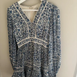 Klänning från By Malina - Supersöt kortklänning från By Malina med blått och vitt blommönster, v-ringning och långa ärmar. Klänningen har volangdetaljer och markerad midja för en feminin siluett. Perfekt för sommarens alla tillfällen! Använt en gång, original pris låg ungefär på 3000kr