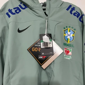 Nike Brasil vindjacka GORE-TEX grön - Snygg grön vindjacka från Nike med Brasil-logga och flera coola tryck på bröst och ärmar. Jackan är tillverkad i GORE-TEX-material, har dragkedja framtill och lång ärm. Perfekt för dig som gillar fotboll och vill ha en sportig look. Priset kan diskuteras.