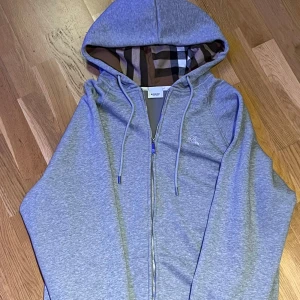 Grå hoodie från Burberry med dragkedja - Säljer en grå hoodie från Burberry med klassiskt rutigt foder i huvan och broderad logga på bröstet. Tröjan har snörning i huvan, hel dragkedja och är tillverkad i 100% bomull. Har ett litet hål inuti men påverkar ingenting.                             TAR EMOT BYTEN
