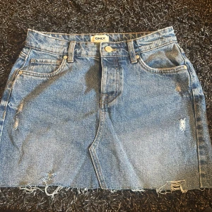 Blå jeanskjol från ONLY med sliten look - Säljer en blå jeanskjol från ONLY med rå kant och slitna detaljer. Kjolen har klassiska fem fickor, knapp och dragkedja framtill samt bälteshällor. Perfekt för dig som gillar en avslappnad och trendig stil.