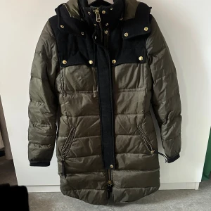 Olivgrön dunjacka från J.Lindeberg - Snygg olivgrön dunjacka från J.Lindeberg med svarta detaljer och guldiga knappar. Jackan är quiltad, har dragkedja framtill och två fickor med dragkedja. Perfekt för kalla dagar och har en längre modell som håller dig varm hela vintern.