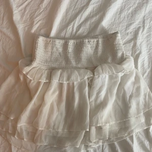 Vit volangkjol från Lindex - Supersöt vit kortkjol från Lindex med bred smock i midjan och dubbla lager volanger. Kjolen är luftig och har en flowy känsla, perfekt för varma dagar. Materialet är lätt och ger en romantisk vibe. Pris kan diskuteras.