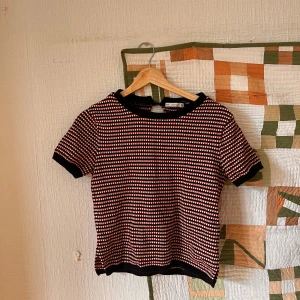 stickad topp från Zara - Säljer en kortärmad stickad topp från Zara med svart, röd och vit randigt mönster. Toppen har ribbade kanter vid hals, ärmslut och nederkant samt en snygg knappdetalj och öppning i nacken. Perfekt för dig som gillar retrovibbar och färg.