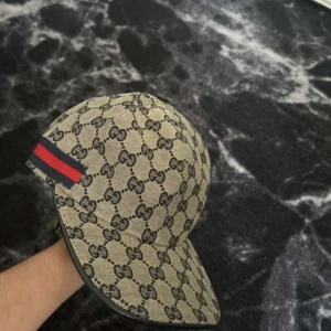 Gucci keps med GG-mönster och röd-blå detalj - Snygg keps från Gucci i beige med klassiskt GG-monogram över hela kepsen. Har en röd och marinblå rand på sidan och justerbar rem baktill i svart läder. Tillverkad i bomullsmix med exklusiv känsla. Perfekt för dig som vill sticka ut med en lyxig accessoar.