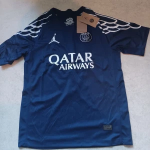 PSG Jordan Dri-Fit fotbollströja Dembele - Snygg marinblå PSG fotbollströja med Jordan-logga, Dri-Fit material och tryckta vita detaljer. Nummer 10 och O. Dembélé på ryggen, Qatar Airways sponsor framtill och Champions League-märke på ärmen. Unikt vingmönster på axlarna. Perfekt för dig som älskar fotboll och streetstyle.
