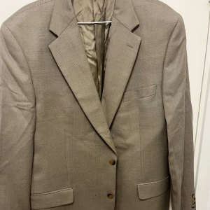 Beige kavaj från Oscar Jacobson - Snygg beige kavaj från Oscar Jacobson med klassisk passform och två knappar framtill. Kavajen har diskreta fickor och är helfodrad med slät insida. Materialet är ull och har en lätt textur som ger en stilren look. Perfekt för dig som vill ha en clean och tidlös stil.