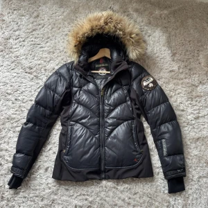 Svart dunjacka med päls från Napapijri - Snygg svart dunjacka från Napapijri med fluffig fuskpäls på huvan och coolt broderat märke på ärmen. Jackan har quiltad design, dragkedja framtill och långa ärmar. Perfekt för kalla dagar och riktigt street i stilen.Skriv snälla om du har några frågor, Kan alltid gå ner i pris!