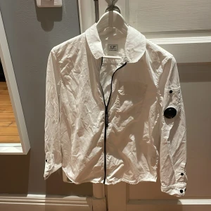 Vit jacka från C.P. Company - Vit jacka från C.P. Company med klassisk krage, svart dragkedja och ikonisk linsdetalj på ärmen. Långärmad modell i bomull med knappdetaljer vid ärmslut och bröstficka. Perfekt för dig som gillar streetwear och stilrena plagg. Riktigt fet till alla årstider. Bara andvända 2 gånger. Och om ni har några mer frågor kan ni bara skriva till mig!