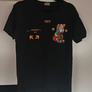 Svart Off-White t-shirt med tryck - Svart t-shirt från Off-White med coolt tryck framtill och stort färgglatt motiv på ryggen. Trycket inkluderar en tecknad figur, sneakers och orangea detaljer. Klassisk passform och rund hals. Perfekt för dig som gillar streetwear och unika prints.
