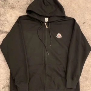 Svart hoodie från Moncler med dragkedja - Svart hoodie från Moncler med huva, snörning och dragkedja framtill. Klassisk logga på bröstet och två fickor. Perfekt för dig som gillar en clean och stilren look. Ribbade muddar vid ärmslut och nederkant. Xl men är liten i storlek sitter som L