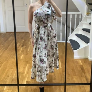 Blommig omlottklänning  - Säljer en superfin långklänning med blommigt mönster. Klänningen har oneshoulder, omlott och ett matchande bälte i midjan. Helt ny! 