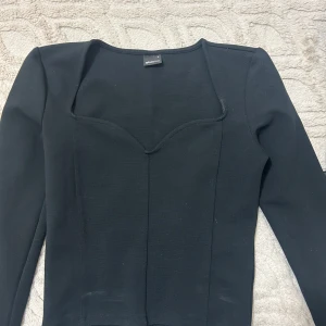 Svart långärmad korsettopp Gina Tricot - Snygg svart korsettinspirerad topp från Gina Tricot med markerade sömmar och lång ärm. Toppen har en unik sweetheart-urringning som ger en cool och trendig look. Perfekt att styla med jeans eller kjol för en modern vibe.