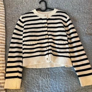 Randig kofta från Gina Tricot - Säljer en snygg kofta från Gina Tricot i svart och vit med breda ränder. Koftan har guldfärgade knappar framtill, två små fickor och ribbade muddar vid ärmslut och nederkant. Materialet är stickad bomull och modellen är normal i passningen.