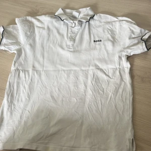 Vit kortärmad BOSS t-shirt med krage - Vit t-shirt från BOSS med klassisk pikékrage och svarta detaljer på krage och ärmslut. Kortärmad modell med knappar vid halsen och diskret BOSS-logga på bröstet. Tillverkad i mjuk bomull som känns skön mot huden.