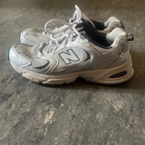 New Balance 530 sneakers vit/silver - Säljer ett par New Balance 530 sneakers i storlek 38.5. Skorna är vita med silverdetaljer och har mesh-överdel för extra ventilation. Klassisk N-logga på sidan, snörning och platt sula. Insidan är fodrad i mörkblått tyg. 