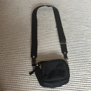 Svart axelväska från H&M - Svart axelväska från H&M med bred justerbar rem och silverfärgade detaljer. Väskan har två fack med dragkedja och en stilren, enkel design. Perfekt storlek för mobil, plånbok och nycklar. Materialet är mjuk polyester.