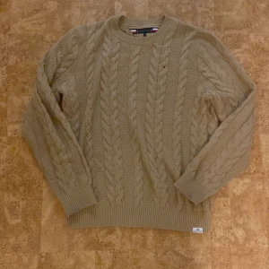 Beige kabelstickad tröja Tommy Hilfiger - Snygg beige kabelstickad tröja från Tommy Hilfiger med klassiskt mönster och diskret logga på bröstet. Rund hals och ribbade muddar vid ärmslut och nederkant. Perfekt för dig som gillar stilrena och tidlösa plagg med lite preppy känsla.