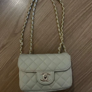 Krämvit/beige quiltad axelväska  - Säljer en stilren vit axelväska med klassiskt quiltat mönster och CC-logga i metall framtill. Väskan har en snygg kedjerem med gulddetaljer och är tillverkad i skinn. Perfekt storlek för det viktigaste och ger en lyxig känsla. Pris går att diskutera.