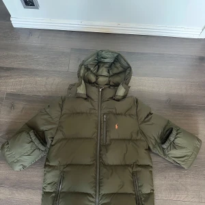 Olivgrön dunjacka ifrån Polo Ralph Lauren - Snygg olivgrön dunjacka  från Polo Ralph Lauren med huva och dragkedja framtill. Jackan har två sidofickor med dragkedja och klassisk Polo-logga på bröstet. Perfekt för kalla dagar med sin vadderade design och stilrena look. Jackan är i storlek XL i barn storlekar vilket motsvarar en S, jackan är väldigt skön och perfekt nu till höst och vinter🙌 Ny pris 3239, priset är prutat och klart hittar du billigare köp den🙂