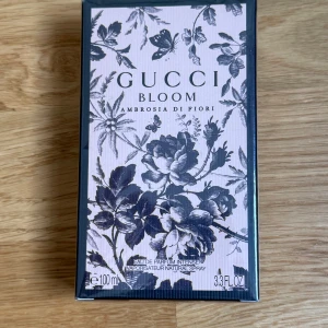 Gucci Bloom Ambrosia di Fiori 100ml - Gucci Bloom Ambrosia di Fiori Eau de Parfum Intense i 100 ml. Lyxig parfym i elegant kartong med blommigt svartvitt mönster och rosa bakgrund. Förpackningen har klassisk rektangulär form och är tillverkad av kartong.