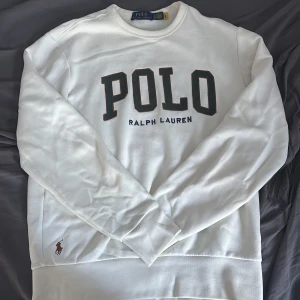 Vit sweatshirt från Polo Ralph Lauren - Vit sweatshirt från Polo Ralph Lauren med stor POLO-text och broderad logga i brunt vid nederkant