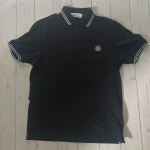 Svart Stone Island pikétröja S - Svart pikétröja från Stone Island med vit piping på krage och ärmslut. Klassisk polokrage med två knappar och ikonisk logopatch på bröstet. Slim fit-modell i mjuk bomull, perfekt för en clean och stilren look.