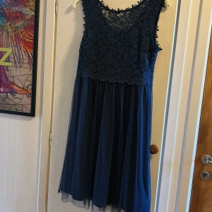 Marinblå spetsklänning med tyllkjol - Supersnygg marinblå klänning med ärmlös design, överdel i blommig spets och kjol i flera lager tyll. Perfekt för dig som vill ha en elegant look med romantiska detaljer och v-ringad rygg. Dragkedja bak för enkel påklädning.