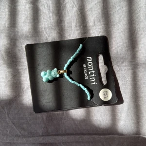 Turkost pärlhalsband från Montini - Snyggt halsband från Montini med små turkosa pärlor och ett hängsmycke i form av en blågrön björn. Kedjan har guldfärgade detaljer och lås. Perfekt accessoar för att ge din outfit en färgklick.