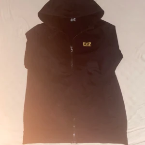 Svart väst från EA7 Emporio Armani - Svart zip up med huva från EA7 Emporio Armani. Tjocktröjan  har dragkedja framtill och en gul logga på bröstet. Tillverkad i mjukt material som känns skönt mot huden. Perfekt för en sportig och stilren look. Tyvärr är zippern sönder men går endå att settas på som en normal hoddie eller tröja inget som märks när man har på den 