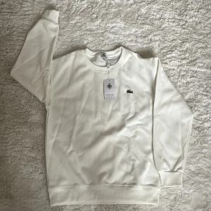 Vit sweatshirt från Lacoste - Clean och stilren vit sweatshirt från Lacoste med klassisk rund hals och broderad krokodil-logga på bröstet. Tröjan har långa ärmar och ribbade muddar vid ärmslut och nederkant. Perfekt för en chill och snygg look.