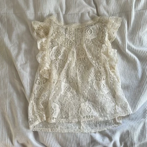 Vit spetsblus med volangärmar - Superfin vit blus i spets med romantiskt mönster och volangdetaljer på ärmarna. Blusen är ärmlös och har en lätt transparent look, perfekt för att skapa en söt och trendig outfit. Passar dig som gillar feminina detaljer och en luftig känsla.