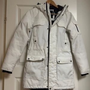 Vit damjacka från Everest - Säljer en vit damjacka från Everest med huva och flera praktiska fickor framtill. Jackan har dragkedja och knappar, samt lång ärm med svart detalj vid ärmslut. Perfekt för kyliga dagar. Har använts 1-2 ggr. Få fläckar finns men det försvinner säkerligen med tvätten. 