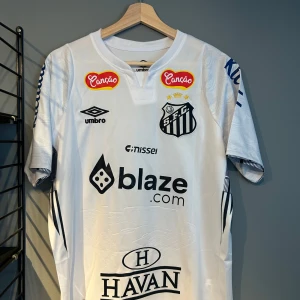 Vit Santos FC Neymar Jr fotbollströja - Santos FC matchtröja i vitt med svarta detaljer och flera sponsorer. Neymar Jr och nummer 10 tryckt på ryggen. Klassisk V-ringning och korta ärmar. Perfekt för fans av brasiliansk fotboll. Helt ny i storlek m en replika 