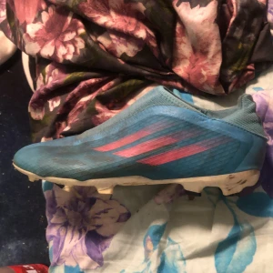 Blåa fotbollsskor från Adidas - Säljer ett par blåa fotbollsskor från Adidas med rosa ränder på sidan. Skorna har en strömlinjeformad design utan snörning och är gjorda i syntetmaterial. Sulan har fasta dobbar.