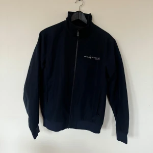 Marinblå vindjacka från Sail Racing - Säljer en marinblå vindjacka från Sail Racing med dragkedja framtill och hög krage. Jackan har logga på bröstet och är tillverkad i ett vindtätt material. Använd fåtal gånger.
