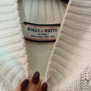 Vit stickad väst från Holly & Whyte - Säljer en vit stickad väst från Holly & Whyte by Lindex. Västen har grovstickad struktur med kabelstickat mönster och bred ribbad krage. Perfekt att styla över en skjorta eller långärmad topp.