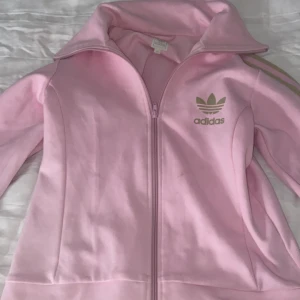 Rosa Adidas zipjacka med guldiga ränder - Säljer en ljusrosa Adidas jacka med dragkedja och guldiga detaljer. Klassiska tre ränder längs ärmarna och Adidas-logga på bröstet. Jackan har hög krage och är långärmad, perfekt för en sportig och trendig look.