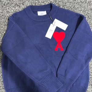 Blå stickad tröja från Ami - Säljer en blå stickad tröja från Ami med rund halsringning och ett stort rött hjärta med bokstaven A broderat på bröstet. Tröjan har långa ärmar och ribbade muddar vid ärmslut och nederkant. Perfekt för dig som vill ha något stilrent men ändå unikt.