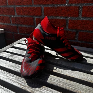 Adidas Predator fotbollsskor i rött och svart - Säljer ett par Adidas Predator fotbollsskor i rött och svart med snörning. Skorna har en smidig och sportig design med röda detaljer och mönster på ovandelen. Perfekta för dig som vill sticka ut på planen!