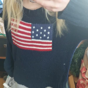 Mörkblå stickad tröja med amerikansk flagga - Säljer en mörkblå stickad tröja med en stor amerikansk flagga på bröstet. Tröjan har rund halsringning och långa ärmar. Perfekt för dig som gillar klassisk och avslappnad stil.