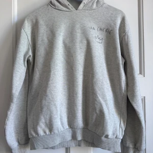 Grå hoodie med tryck från Gina Tricot - Säljer en ljusgrå hoodie från Gina Tricot med texten 'MA CHÉRIE' och en krona tryckt både fram och bak. Hoodien har långa ärmar, mjuk insida och ribbade muddar vid ärmslut och nederkant. Perfekt för en chill och avslappnad stil.