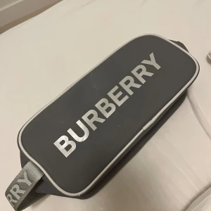 Grå sminkväska från Burberry - Snygg grå sminkväska från Burberry med vit logotyp framtill och vita detaljer längs kanterna. Får plats med MASSA smink och har ett litet fack i sig för att lägga mindre saker i. Har  använt 2 gånger så den har fått lite puder på sig men det går bort med en servett hann bara inte ta bort innan jag tog bilden. 
