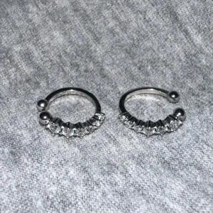 Silverfärgade örhängen med stenar - Säljer ett par snygga silverfärgade örhängen med glittrande stenar framtill. De har en öppen design med kulor i ändarna och är till för att användas där man inte har hål. 