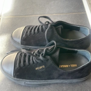 Svarta sneakers från Axel Arigato - Säljer ett par svarta sneakers från Axel Arigato med ovandel i mocka och tåparti i slätt material. Skorna har snörning och platt sula, samt diskret logga i guld på sidan. Perfekta för en stilren och enkel look.