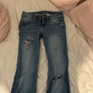 Blå jeans med slitningar - Säljer ett par blå jeans med slitna detaljer på båda ben, de är raka ben med normal passform. Perfekta för en avslappnad och trendig look. Midjemått 35cm