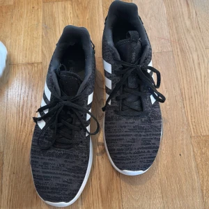 Svarta Adidas sneakers med vita ränder - Säljer ett par svarta Adidas sneakers med klassiska vita ränder på sidorna. Skorna har svart snörning, rund tå och platt vit sula. Ovandelen har ett diskret mönster i svart och grått. Perfekta för vardagsbruk eller träning. Storlek 43 1/3. Använda fåtal gånger och är i fint skick