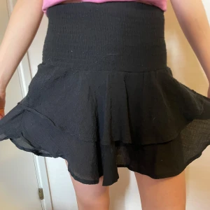 Svart volangkjol med resårmidja🖤 - Säljer en svart kortkjol med bred smockad resårmidja och dubbla volanglager. Kjolen har en luftig och flowy känsla. Man kan ha strumpbyxor under eller inte och den är perfekt för både våren och sommaren😜