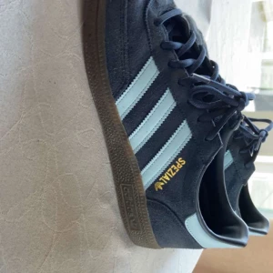 Adidas Spezial sneakers i mörkblå mocka - Säljer ett par Adidas Spezial sneakers i mörkblå mocka med ljusblå ränder och klassisk gummisula. Skorna har snörning och den ikoniska Spezial-loggan i guld på sidan. Perfekta för dig som gillar retrostil och streetwear.